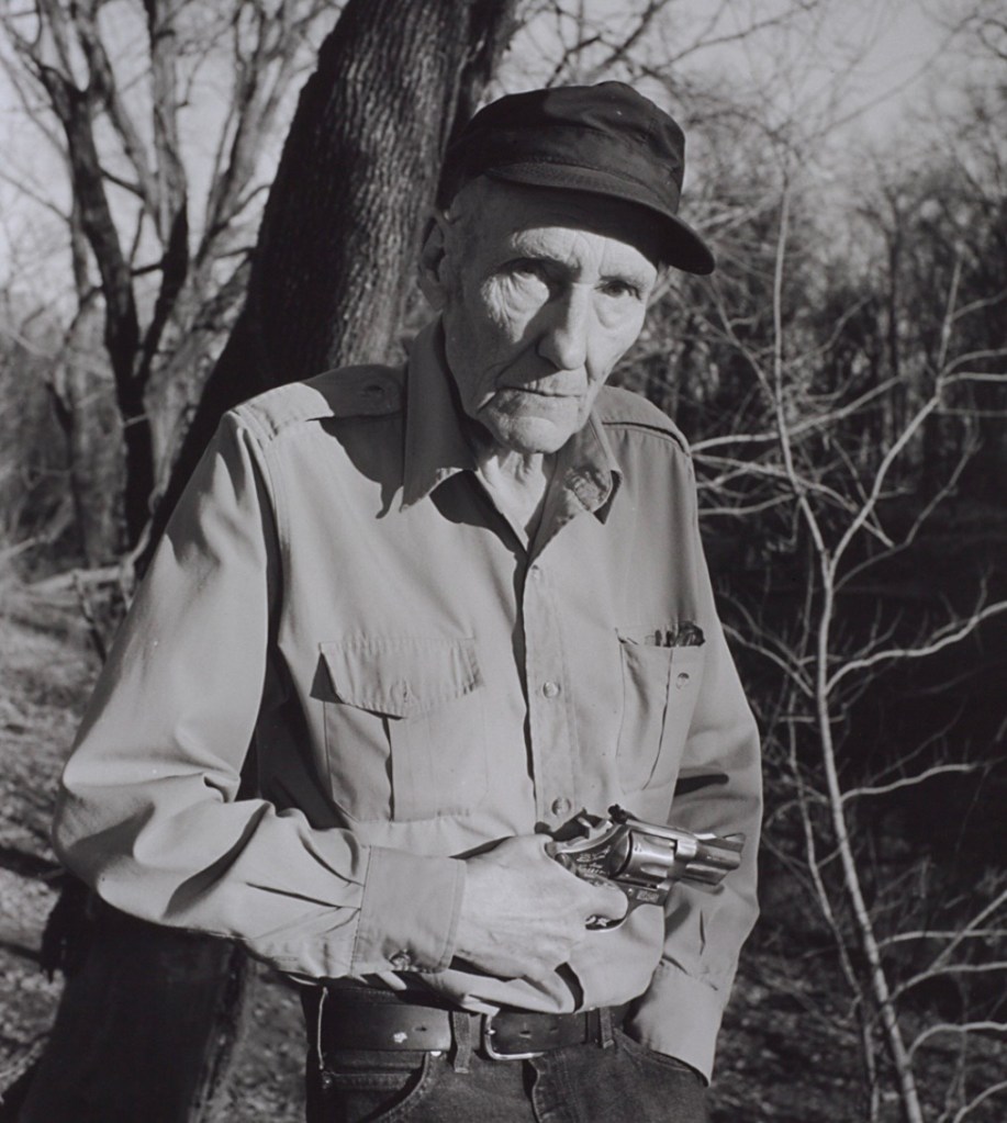 An elderly man holding a revolver, standing outdoors near bare trees in black and white.
