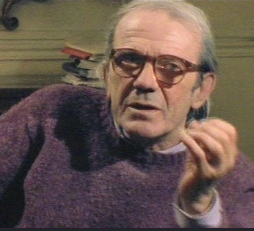 A man with gray hair and glasses is speaking while sitting and gesturing with his hand. He is wearing a purple sweater against a blurred background of books.