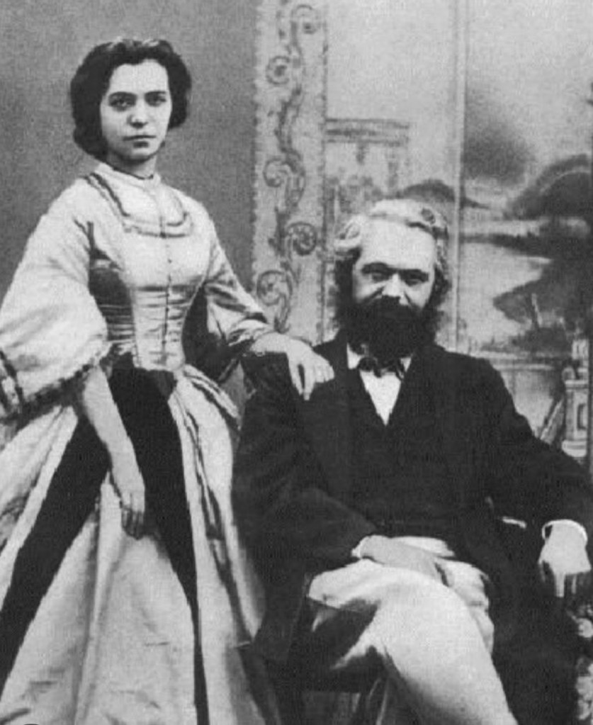 A vintage black and white photograph of a man seated and a woman standing beside him. The man has a beard and mustache, wearing a suit, while the woman is dressed in a long, elegant gown.