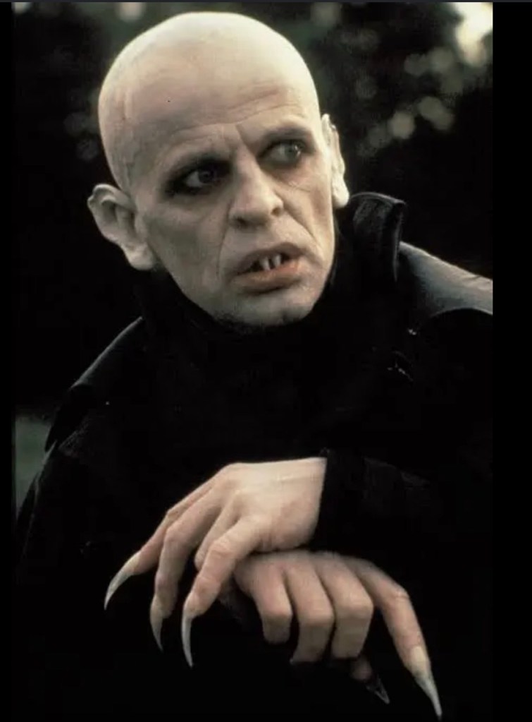 A character with a bald head and pronounced features, wearing dark clothing, with long claws on their fingers, set against a blurred dark background.